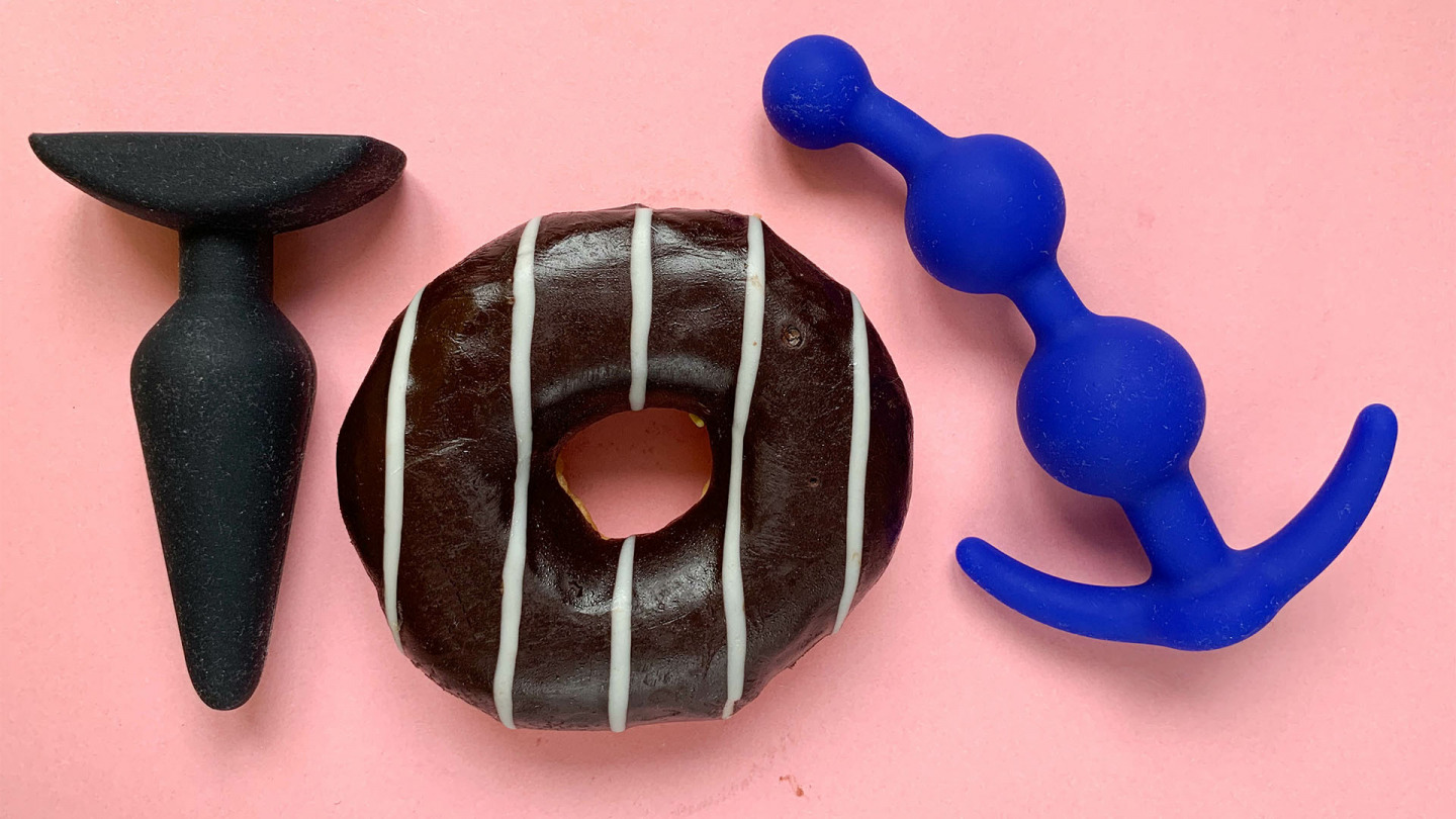 Imagem de dois plugs anais, um liso, outro ondulado ao lado de um donut coberto com chocolate e fios de chocolate branco